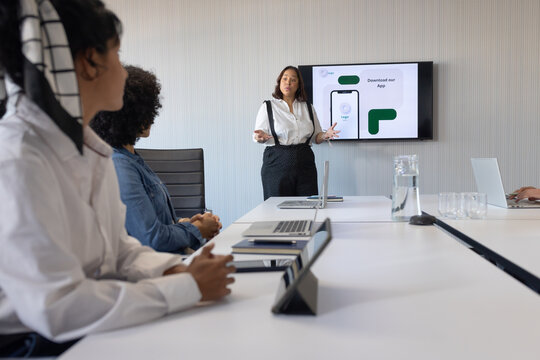 Female colleagues seated in conference room, presenter standing gesturing at monitor showing app