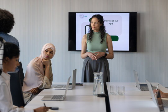 Diverse female coworkers sitting in meeting room, presenter standing, presenting smartphone slide