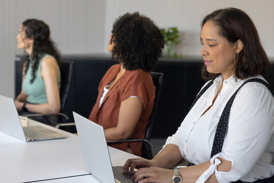 African American women typing at conference table with laptops, right wearing suspenders