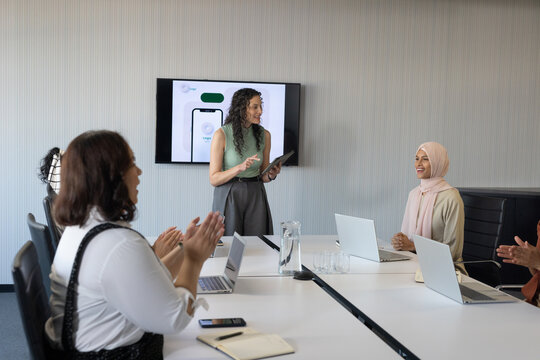 Diverse female coworkers presenting app mockup at conference table with laptops, headscarf visible