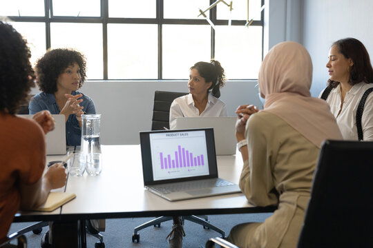 Diverse female coworkers discussing product sales on laptop with purple chart at meeting table