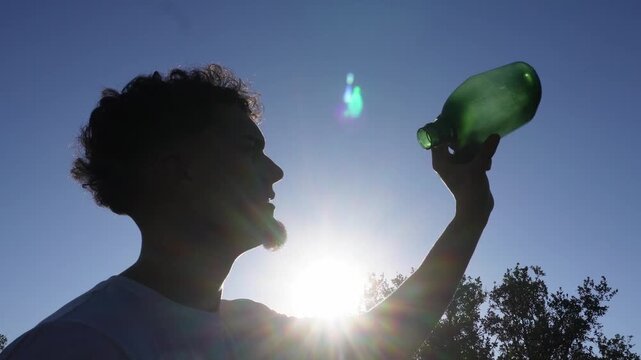 Silhouette of a young man with curly hair drinking water from a green glass bottle against the bright sun. Concept of thirst and dehydration