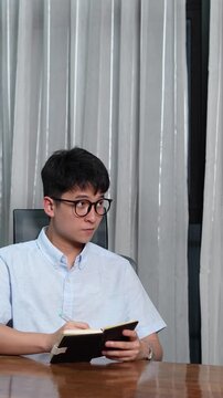 Asian young man writing in a notebook at a desk, standing up to greet a faceless client with a respectful two-handed handshake, business meeting and job interview concept, vertical video
