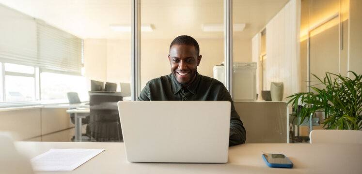 Web banner of happy african business man in 30s smiling at camera behind laptop at office desk, black consultant working online with paper document and mobile phone, corporate staff. Copy space.