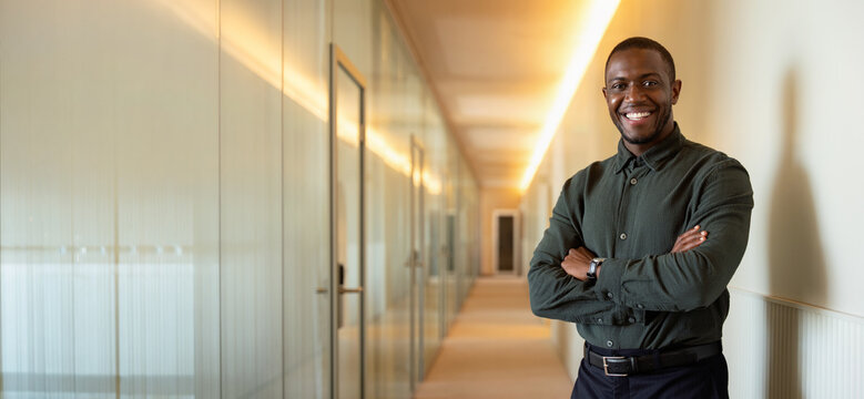 Web banner of black business man in 30s standing at modern office corridor, smiling african CEO and company owner, executive portrait for corporate website, blog or landing page. Copy space.