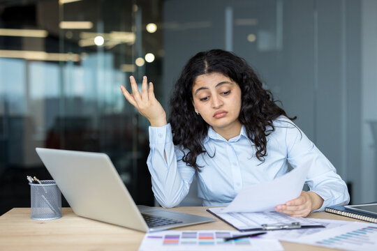 Stressed Businesswoman Working On Laptop And Documents In Office While Feeling Overwhelmed By Financial Analysis Tasks. A Professional Female Manager Reading Paper Documents At Her Desk.