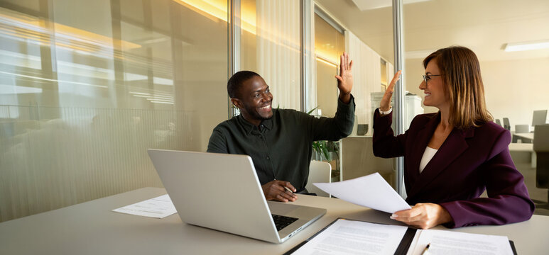 Web banner of two happy business colleagues giving high five at office desk with laptop and paper documents, african man in 30s celebrating deal with european woman in 40s, teamwork. Copy space.