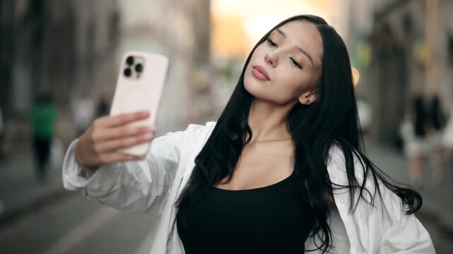 Beautiful attractive young woman taking selfie on smartphone in old European city street. Pretty brunette female posing and holding mobile phone outdoors on blurred urban background