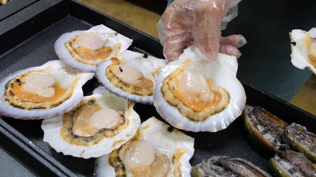 Fresh seafood items including raw scallops and abalone are carefully organized on dark metal trays within a commercial kitchen environment for professional culinary preparation and service operations