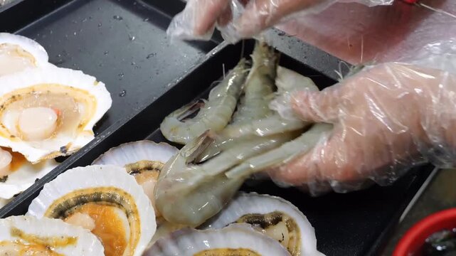 Fresh seafood items including raw shrimp and scallops are carefully organized on dark metal trays within a commercial kitchen environment for professional culinary preparation and service operations
