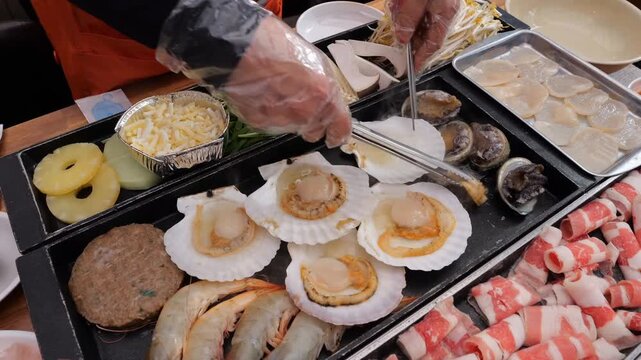 Various proteins including seafood and meats cook on a hot griddle alongside fresh vegetables and herbs, then get transferred into dipping sauces and plates for a meal service