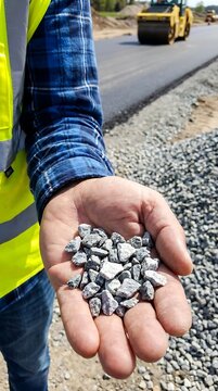 Construction worker holding a handful of crushed grey gravel stones at a road construction site with a heavy steamroller in background
