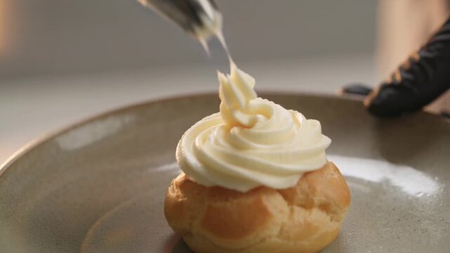 Squeezing piping bag, star nozzle, gloved hand piping rosette onto choux pastry on plate, to finish