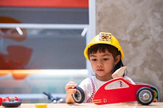 Young girl wearing a safety helmet while repairing a toy vehicle with a wheel, exploring pretend construction play, creativity, problem solving, practical skills, imagination, and career learning.