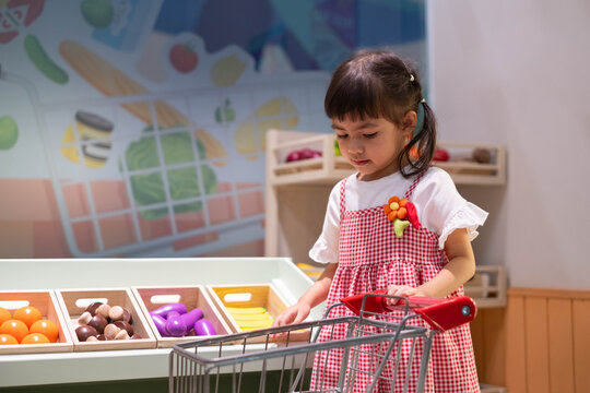 Young girl shopping for colorful toy vegetables in a pretend grocery store, pushing a shopping cart and learning about healthy food choices, nutrition, independence, creativity, and childhood play.