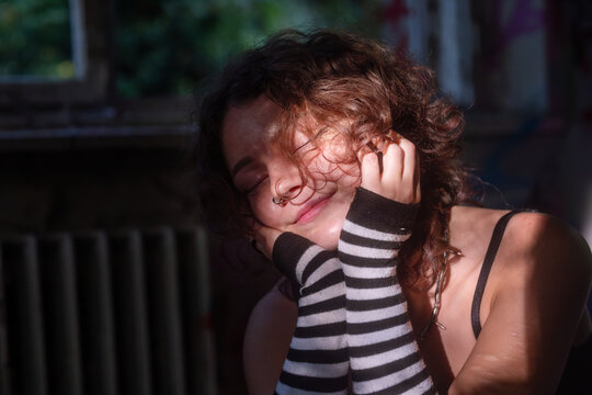 Young woman with curly hair and nose piercings rests her face in striped arm warmers with closed eyes, creating an intimate alternative lifestyle portrait