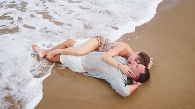 High angle view of man and woman lying embraced on beach while ocean waves gently wash over them, symbolizing companionship, emotional support, trust and comfort in a close loving relationship