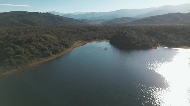 Drone flight tracing a glimmering reservoir bend through hazy hills