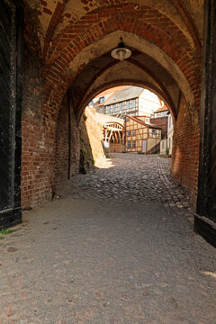 View through the pointed arches of a historic brick gate toward half timbered houses and a cobblestone street, photographed in vertical format.