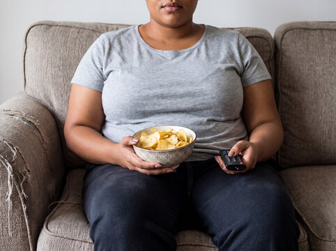 Overweight woman relaxing on couch eating potato chips and watching tv, representing unhealthy lifestyle
