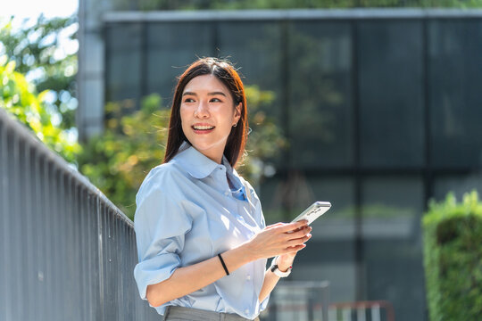 Asian businesswoman leans against a metal railing outside her office and looks away from her smartphone during a short break on a sunny afternoon.