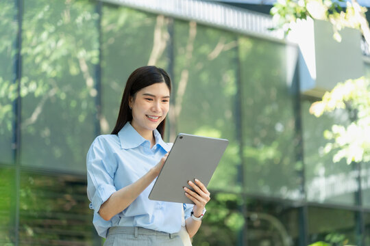 Asian businesswoman in a light blue shirt reads on a digital tablet while standing outdoors in front of a reflective glass office facade.