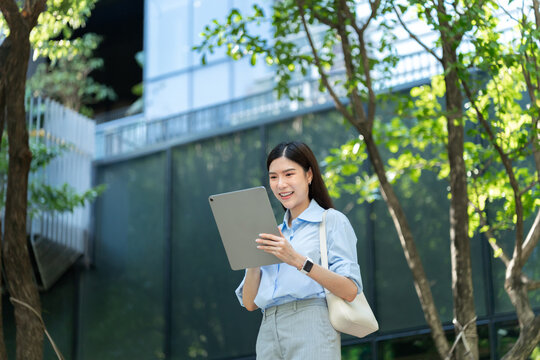Asian businesswoman with a shoulder bag reads on a tablet computer while standing among green trees beside a modern office building.