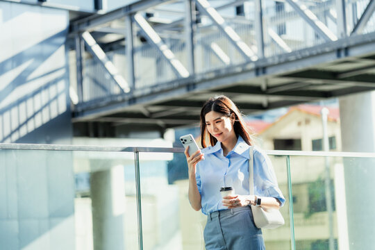 Asian businesswoman walks along an elevated city walkway holding a smartphone and a takeaway coffee cup, with a steel bridge overhead.