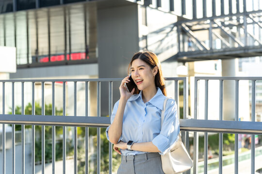 Asian businesswoman talks on her mobile phone at the railing of a city walkway, backlit by warm evening sun in the business district.