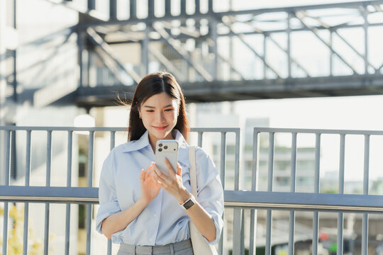 Asian businesswoman stands at the railing of an elevated walkway and types on her smartphone in the business district in bright daylight.
