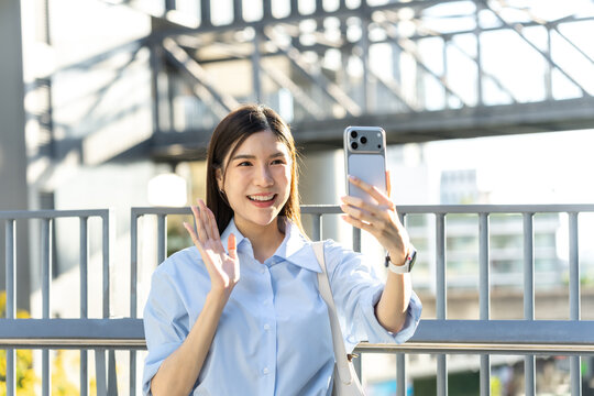 Asian woman raises a hand to greet someone on a mobile video call while standing at the railing of a city bridge in warm afternoon light.