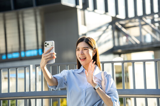 Asian businesswoman holds up her smartphone and waves at the front camera during a video call on an elevated city walkway.