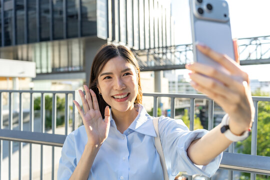 Asian businesswoman stretches out her arm to hold a smartphone for a video call, with city buildings blurred in the background.