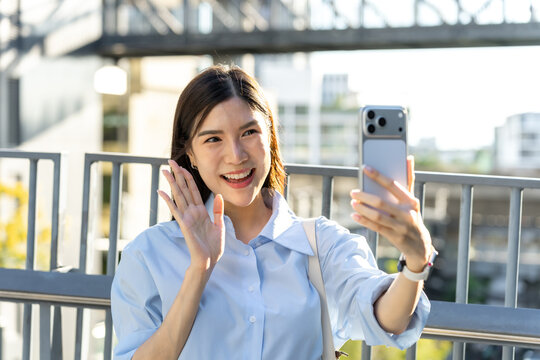 Close view of a smiling asian woman waving at her smartphone front camera during a video chat on an elevated city walkway.