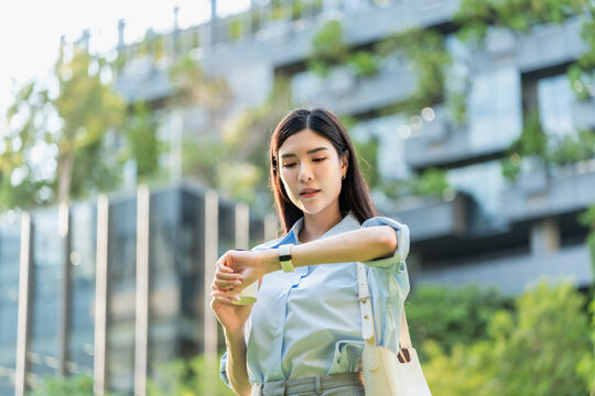 Asian businesswoman checks the time on her smart watch while standing outside an office building with a planted vertical garden.