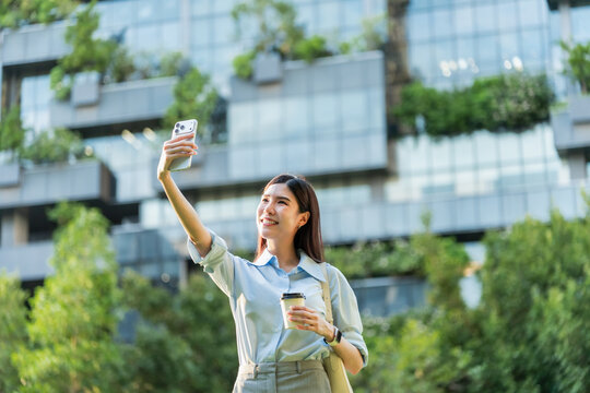 Asian businesswoman raises her smartphone to take a selfie in front of a building covered with green plants, holding a coffee cup.