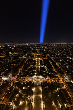 A view from the top of the Eiffel Tower at night, overlooking the city of Paris. The searchlight from the tower extends toward the horizon.