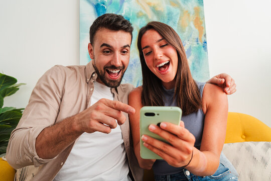 Two smiling people sit close on a couch, sharing a funny moment as a smartphone reveals surprising content. Focus on hands and device, lighting soft, candid mood, and effortless interaction during a
