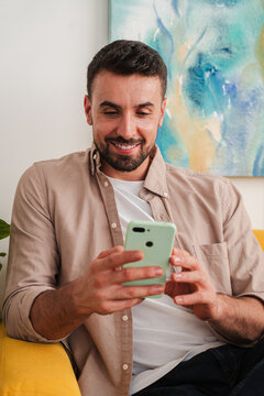 Candid shot of a man focused on a green smartphone, relaxed in a comfortable chair, enjoying a moment of digital interaction. Clean composition highlights concentration and modern tech usage. Calmly.