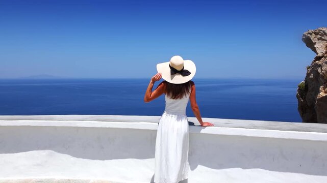 A woman in a white summer dress enjoys the elevated view over the Aegean Sea from a viewpoint at Folegandros Island, Cyclades, Greece