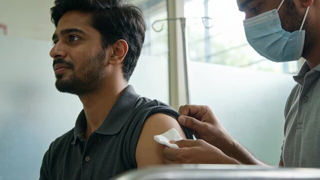 Pulling sleeve up, Indian man dark-polo masked medic wiping deltoid with cotton pad vaccine clinic