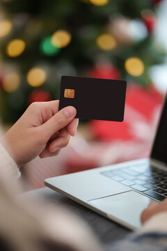 Woman holding a credit card beside a laptop during Christmas, representing online shopping, holiday spending, digital payment, e-commerce checkout, seasonal discounts and festive retail sales.