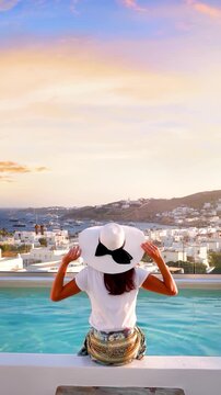 Rear view of a woman sitting by the swimming pool and enjoying the summer sunset view over Mykonos island, Cyclades, Greece