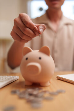 Man inserting a coin into a piggy bank at home with calculator and coins nearby, representing savings, budgeting, financial planning, money management, investment and future security.