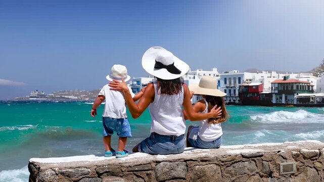 A tourist mother sits with her children on a wall and looks at the town of Mykonos island, Cyclades, Greece, on a windy summer day