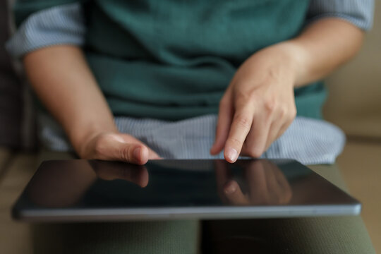 Close-up of a woman using a touchscreen tablet while sitting at sofa, representing digital technology, online access, mobile communication, browsing, smart devices and modern lifestyle.