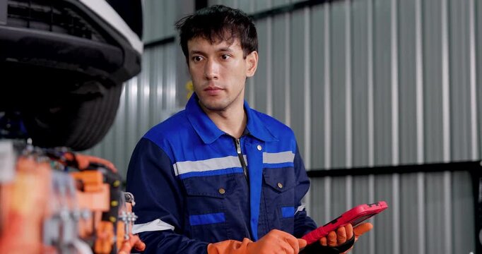 Young Asian male technician reviews EV battery components while using digital tablet in automotive workshop, performing electric vehicle diagnostics, battery inspection, maintenance assessment.