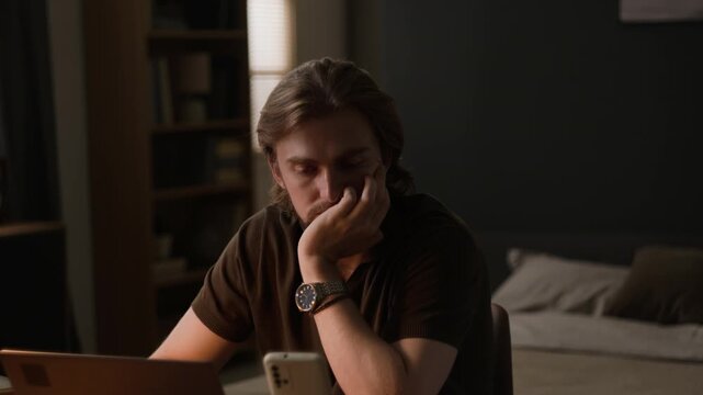 Caucasian young adult man looking exhausted and stressed while working with laptop and smartphone at home. Perfect for projects on remote work fatigue, digital burnout, and mental health awareness