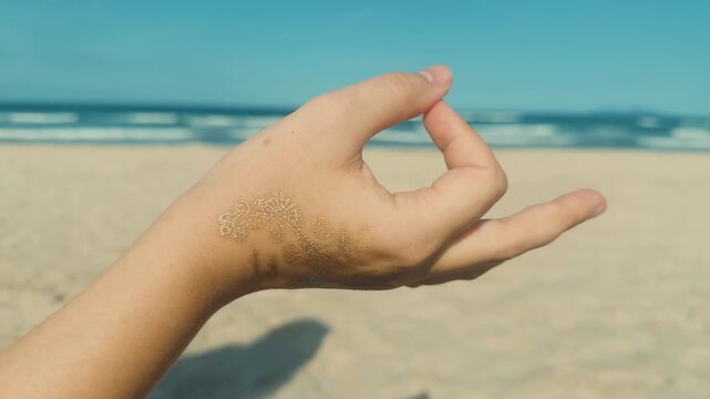 Hand forming a traditional meditation mudra gesture with a decorative temporary metallic tattoo, symbolizing peace and spiritual mindfulness on a sandy beach with the ocean in the background