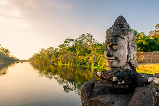 Deva head statue at Angkor Thom South gate, smiling serene Buddha face at sunset in Angkor, ancient moat with water reflections, Siem Reap, Cambodia
